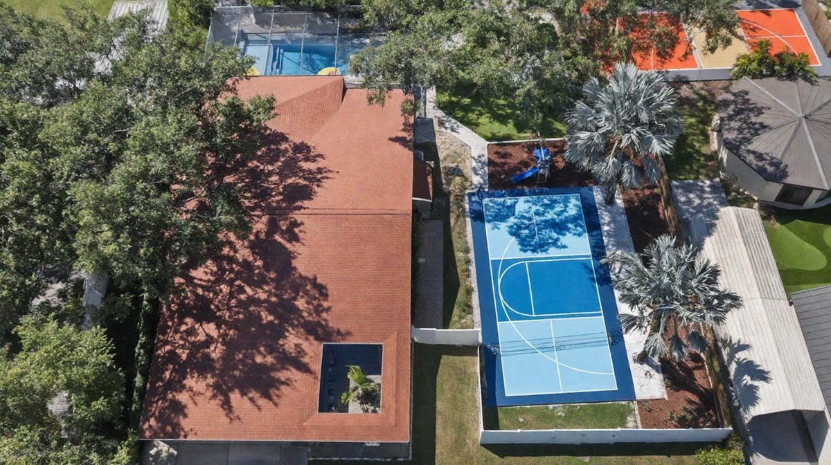 Aerial of a blue basketball court beside a tile-roof home, ringed by palms with a pool and playground nearby