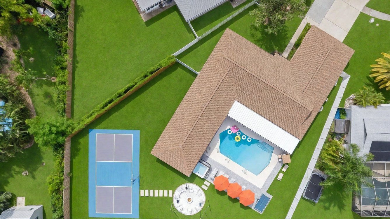 Aerial view of a GR estate: pickleball court, pool with floats, orange umbrellas and a paver fire-pit circle on a manicured lawn