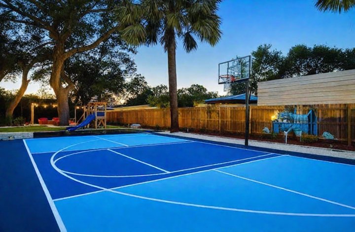 A vivid blue basketball court at dusk with a playground and string lights glowing under palm silhouettes